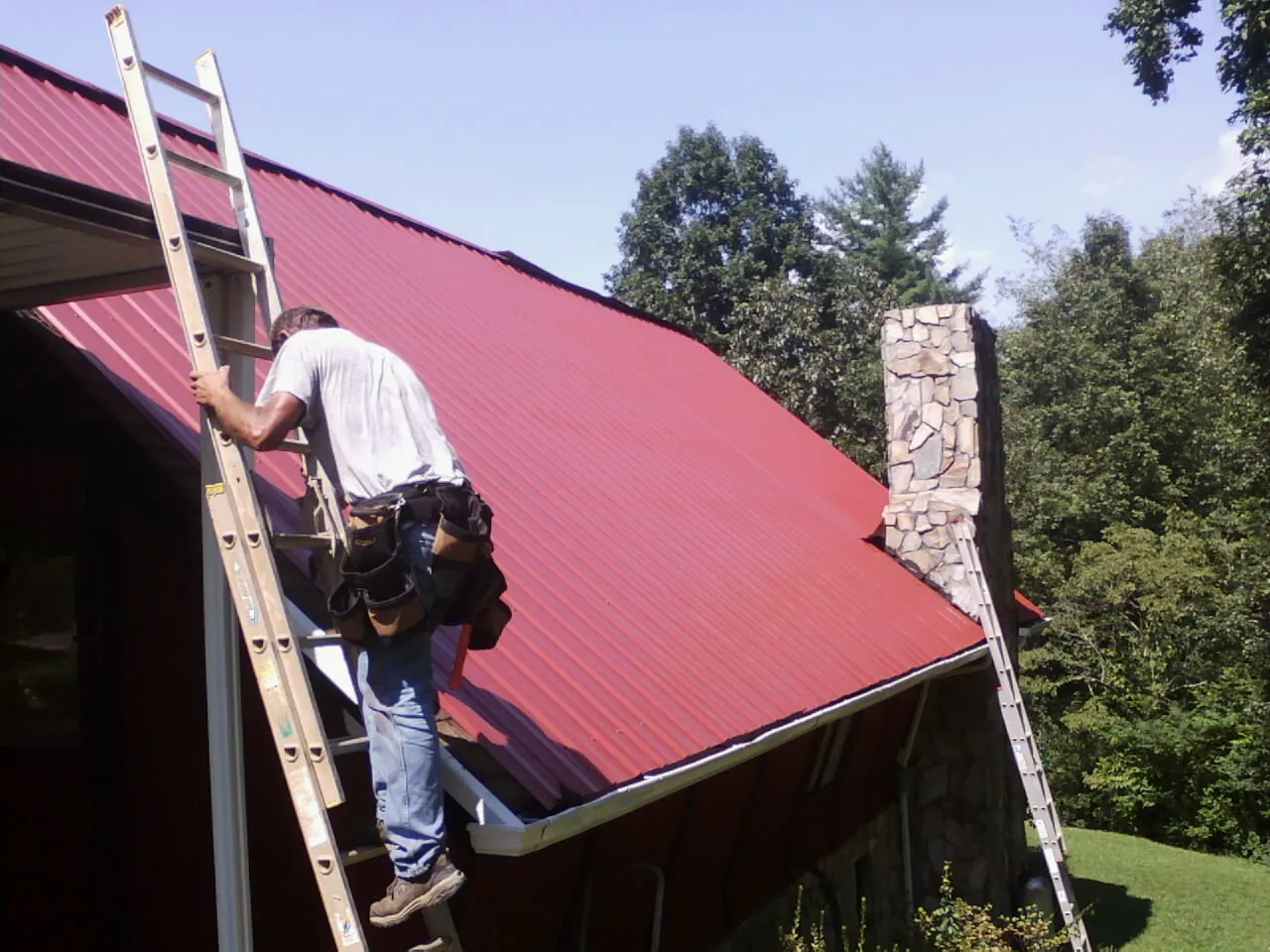 Roofer on a ladder beside a red metal roof and stone chimney — Soffit & Fascia Repair by Roofing Brothers in Park Hills
